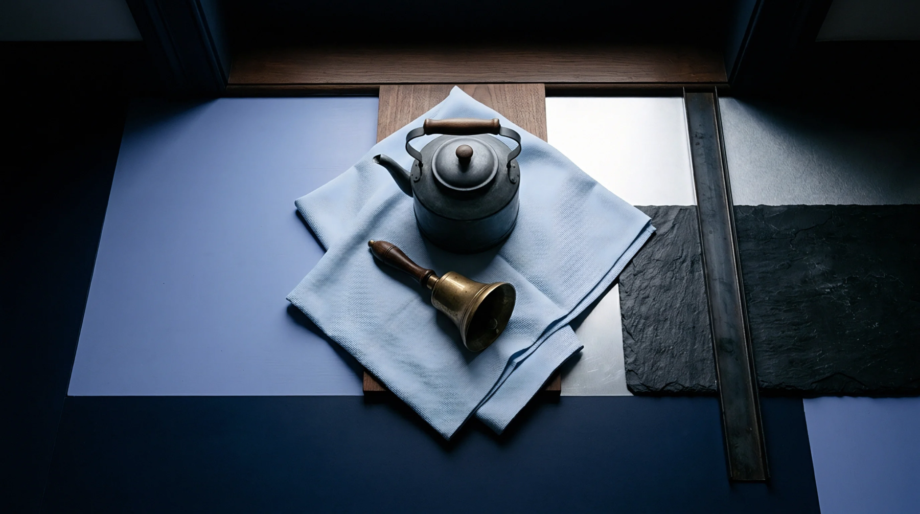An aged metal kettle and a brass bell rest on light blue textile over varied raw material samples and industrial surfaces.