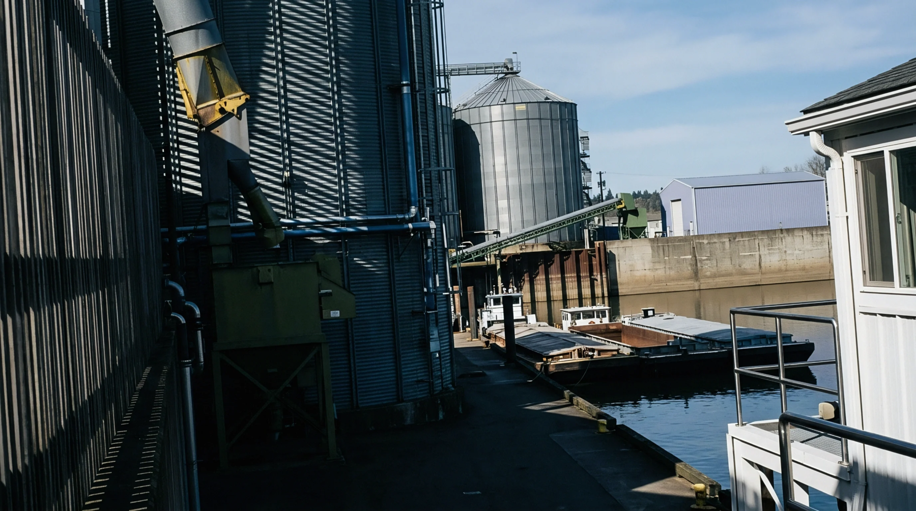 Corrugated steel grain silos and conveyors stand beside docked transport barges on an industrial waterway terminal.