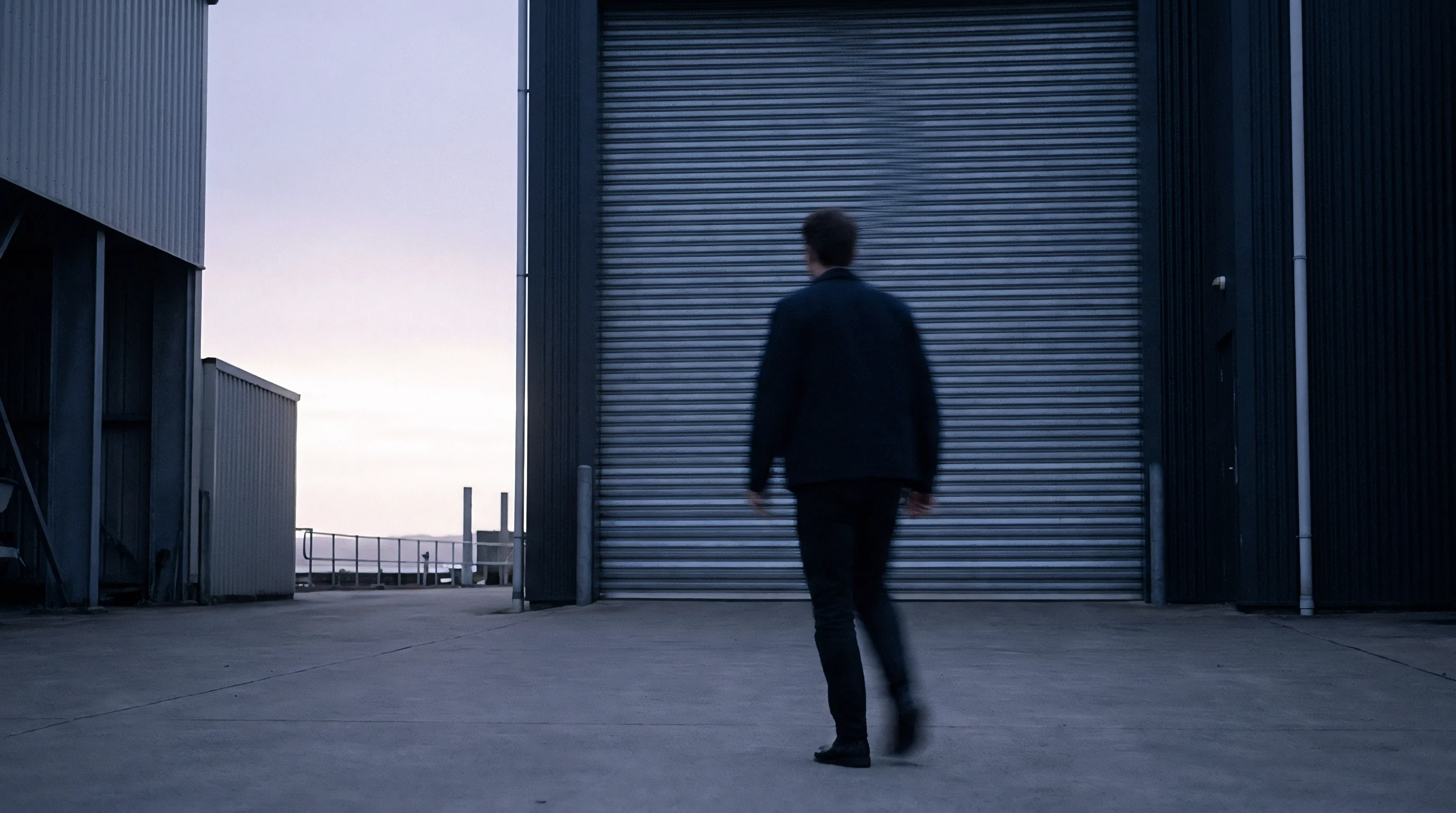 A worker in dark outerwear walks across a concrete courtyard toward a large corrugated metal loading door at an industrial facility.