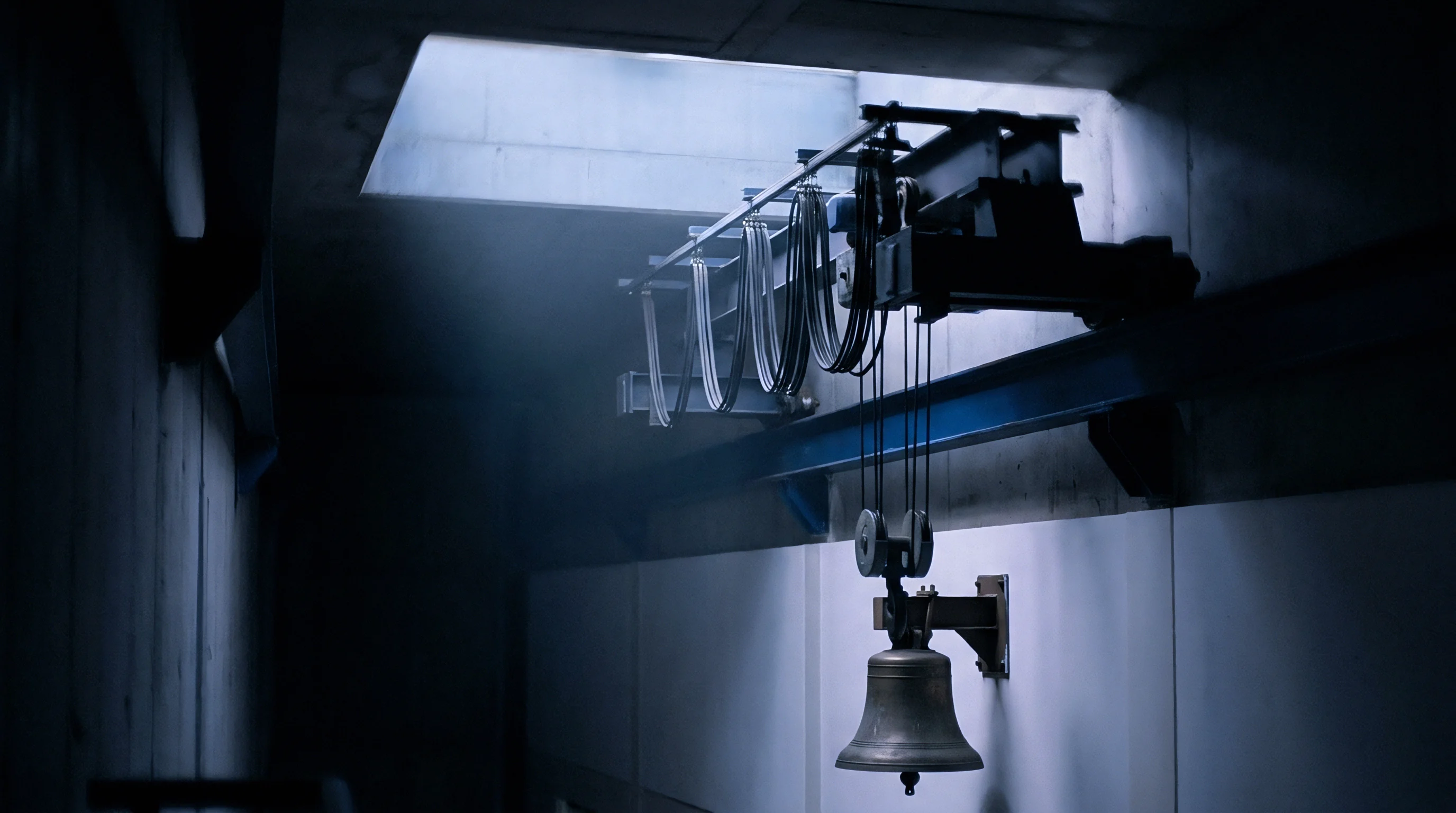 Structural steel beams support an overhead crane assembly with heavy rigging cables above a metal signal bell inside a concrete industrial facility.